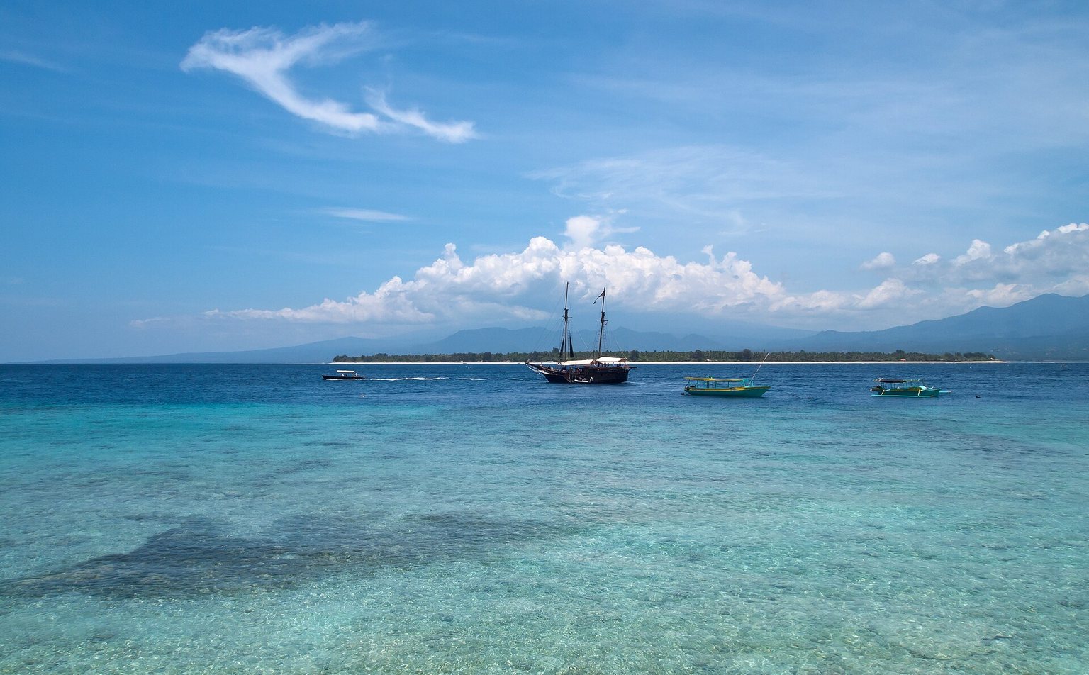 Clear turquoise shallows off Gili Meno with a wooden sailing ship and small boats, and Gili Air low on the horizon