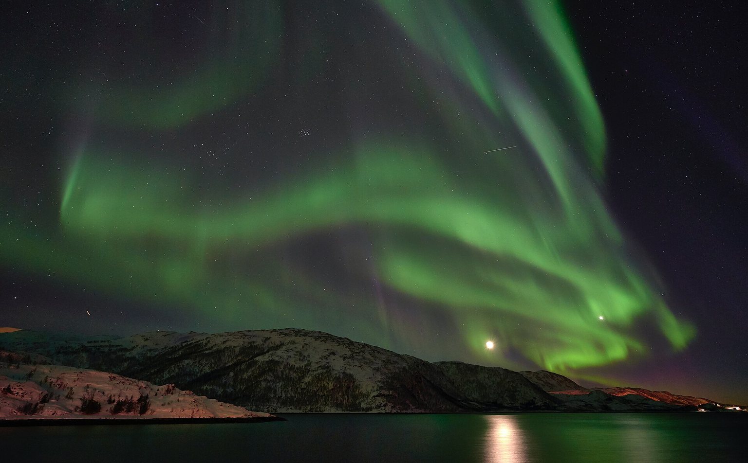 Green aurora arcs over a snowy ridge and a calm fjord at Lauklines on Kvaløya, near Tromsø