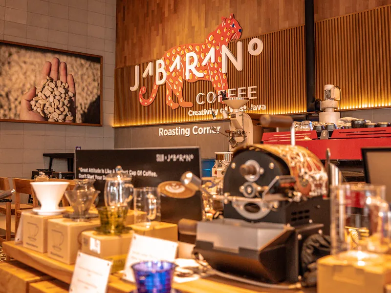 Inside Jabarano Coffee on Jalan Braga, Bandung: a small coffee roaster on a wooden counter below the café’s leopard logo and a Roasting Corner sign